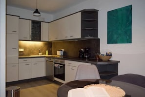 Une cuisine moderne avec des armoires blanches, un four encastré et une machine à café. On y trouve un coin repas avec un canapé de couleur foncée et une petite table. Un tableau abstrait vert est accroché au mur.