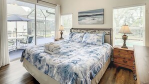A bedroom with a bed, a nightstand, a lamp, a painting, and a view through large windows. - Glenbrook Vacation Pool/Spa Home Near Disney (Clermont)