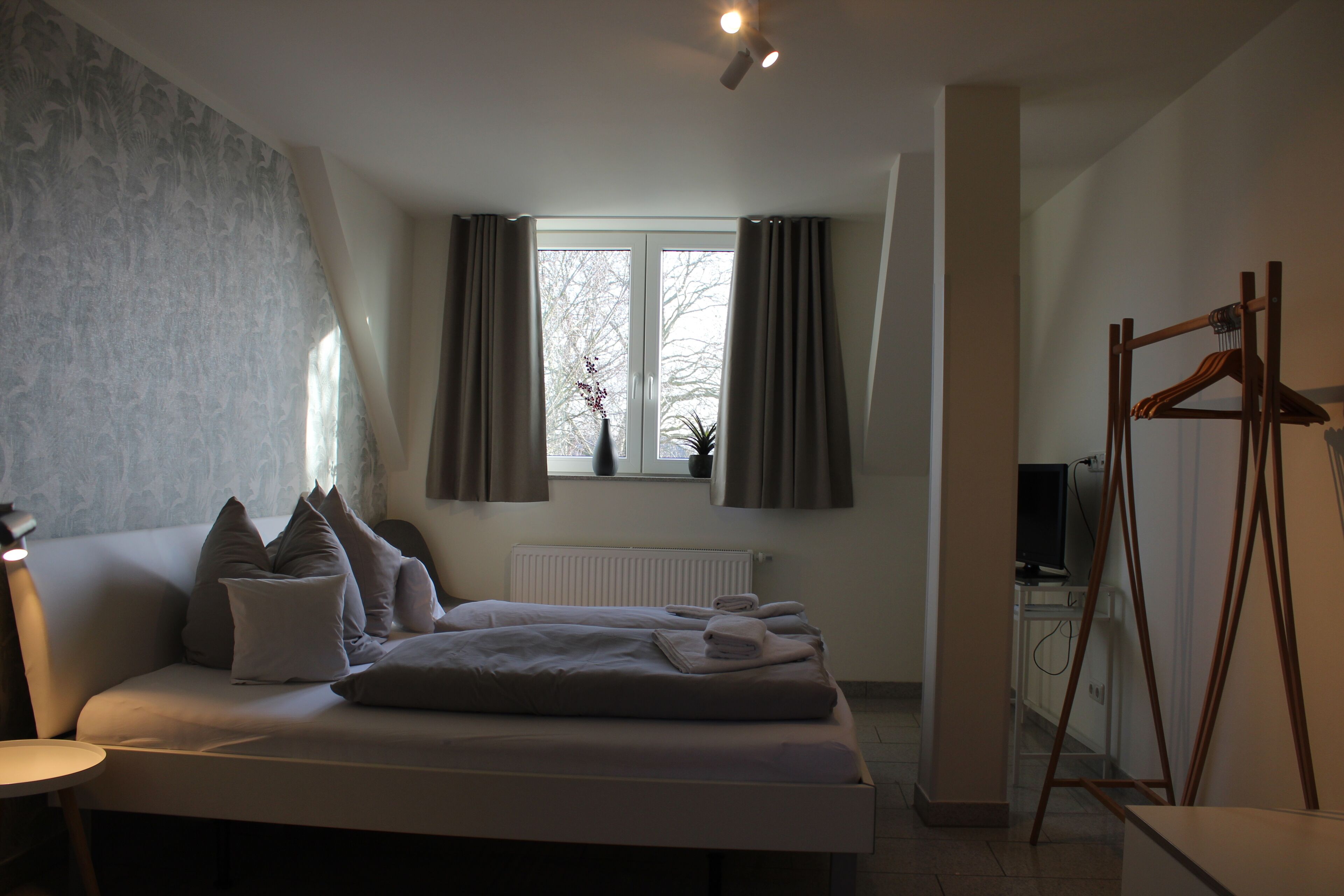 A modern bedroom with a bed, a bedside table, a window with curtains, a radiator, and a TV stand.