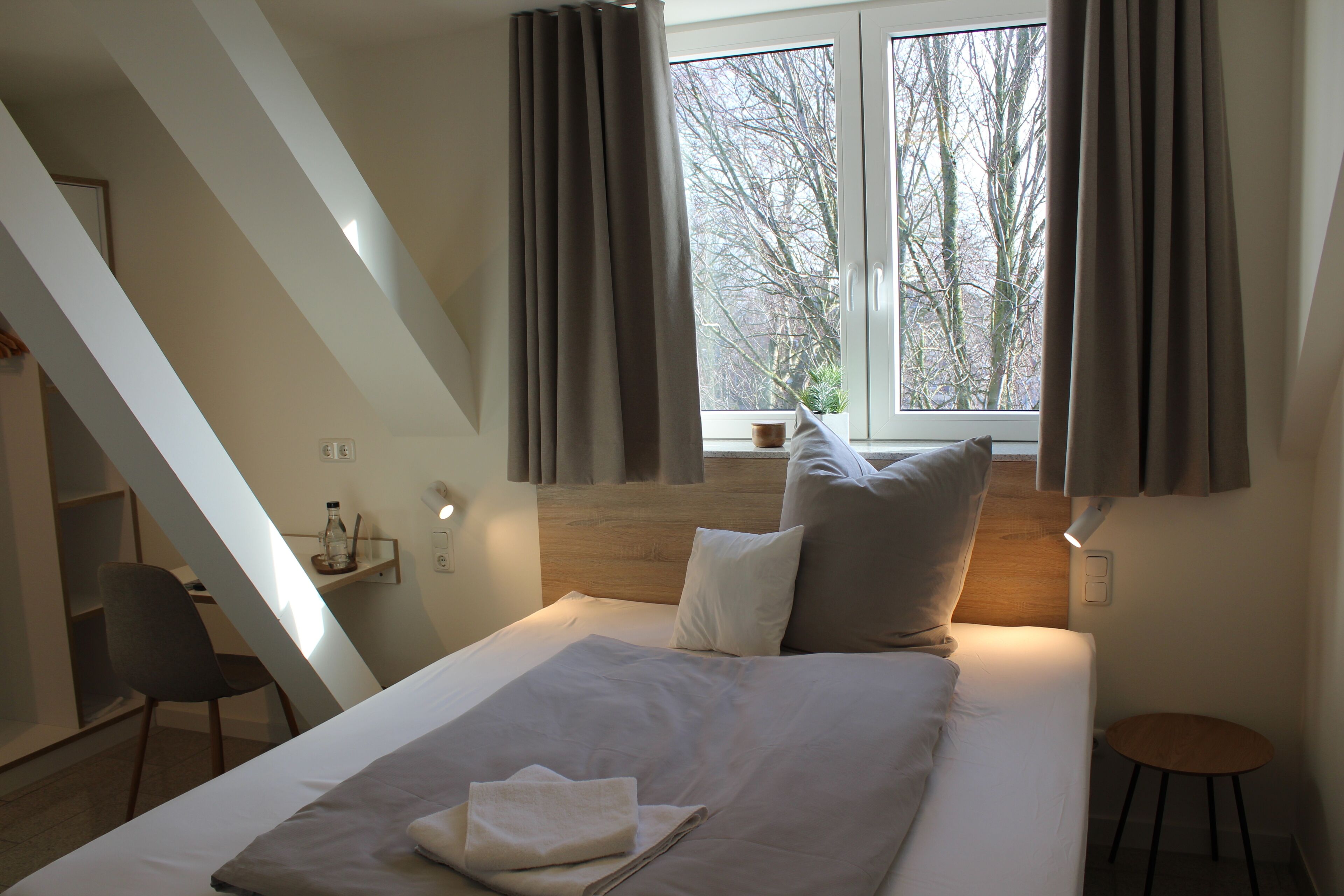 A modern bedroom with a sloped ceiling, a bed with white linens, two pillows, a bedside table, and a window with curtains.