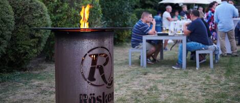 A metal outdoor grill with flames, surrounded by a grassy area and a group of people at a table under an umbrella.