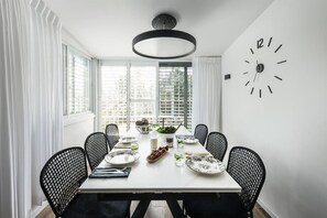 A dining room with a white table, black chairs, a large wall clock, and a window with blinds. - Relax in a Rural Retreat 2BR Apt wGarden (Ramat Hasharon)