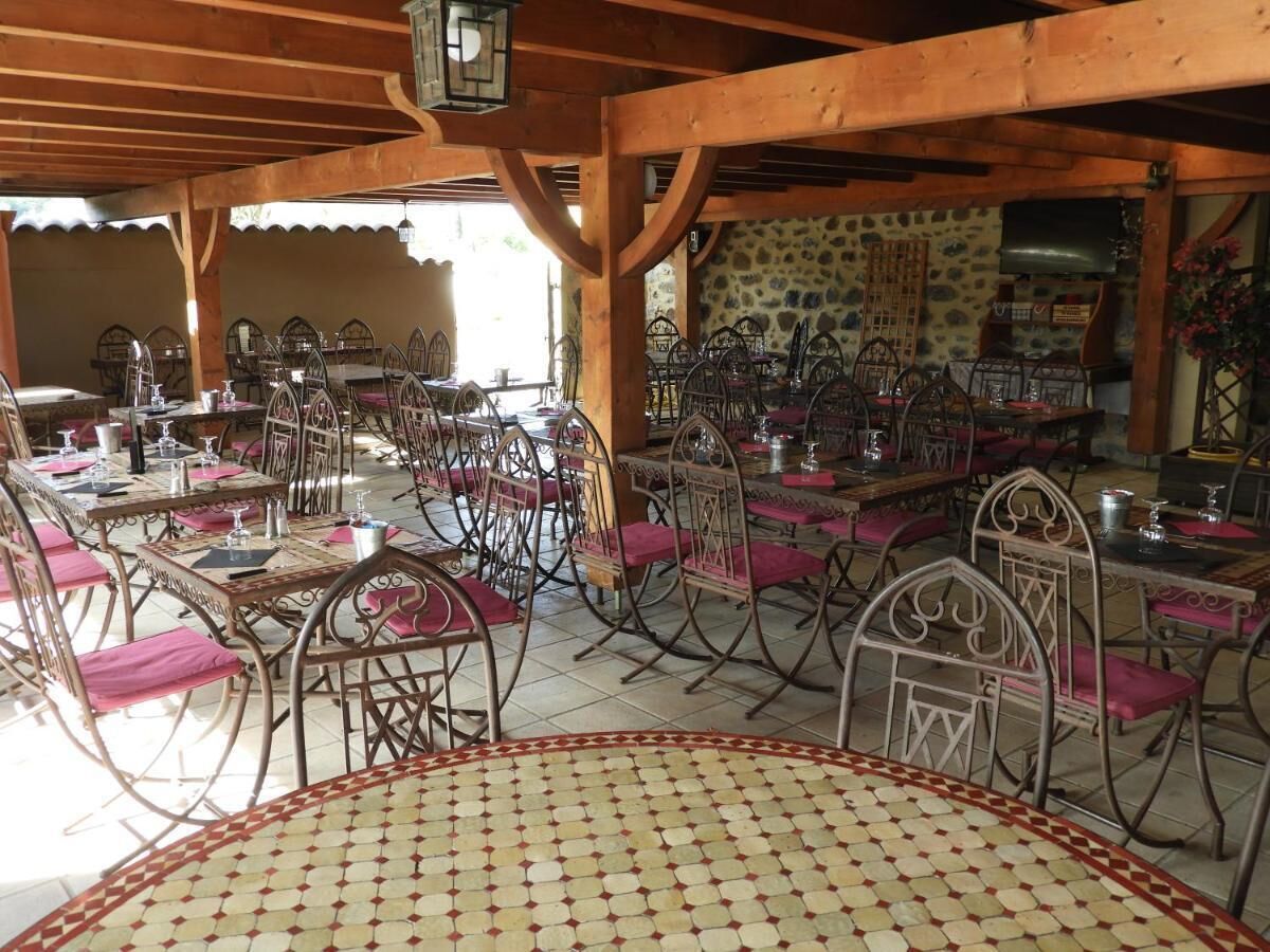 An outdoor dining area with multiple tables and chairs, a stone wall in the background, and a wooden ceiling structure.
