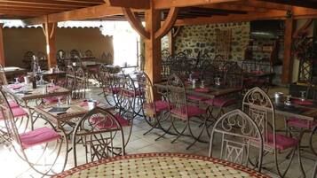 An outdoor dining area with multiple tables and chairs, a stone wall in the background, and a wooden ceiling structure.