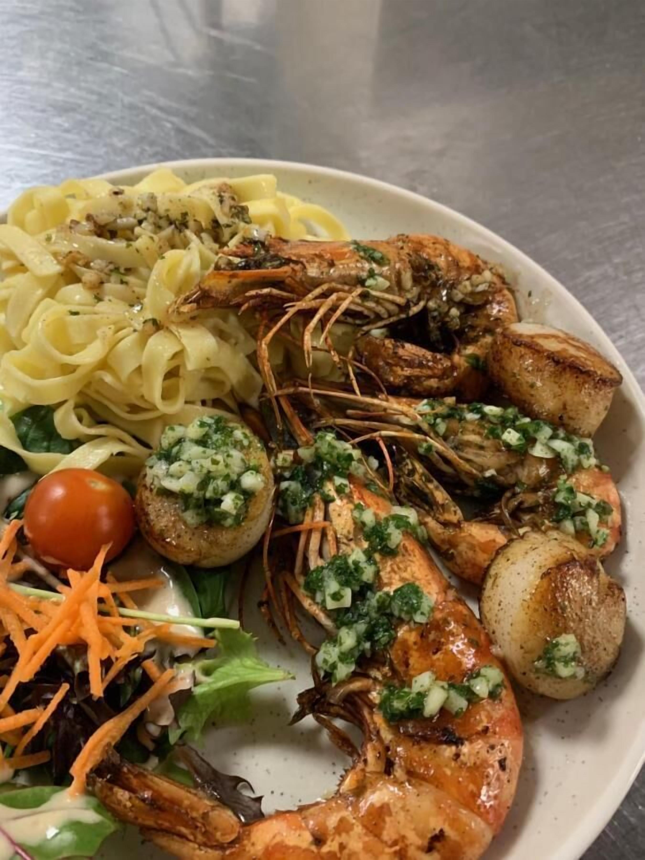 A plate of food including grilled shrimp, scallops, pasta, and a salad with a cherry tomato and shredded carrots.