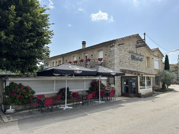 Un edificio de piedra con barra y zona de restaurante, mesas al aire libre con sombrillas y una entrada visible.