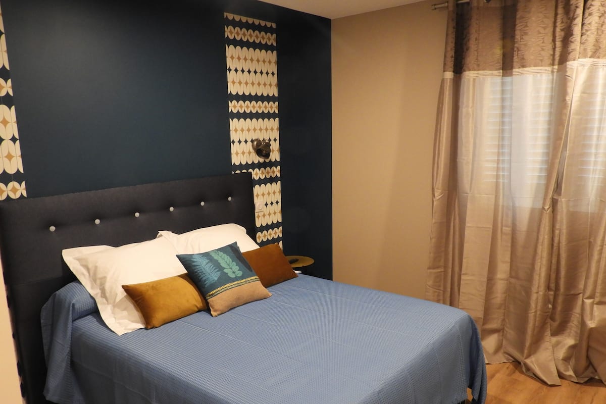 A neatly made bed with a blue bedspread and multiple pillows, a dark headboard, and a wall with a geometric pattern.