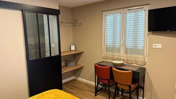 A compact room with a bed, a desk with two chairs, a window with blinds, and a TV mounted on the wall.