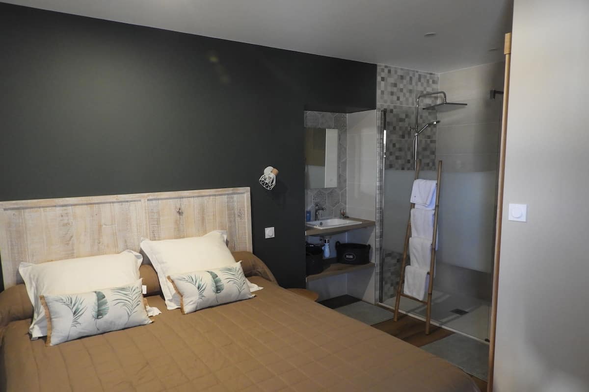 A bedroom with a bed, pillows, a bedside table, a mirror, and a bathroom with a shower and sink.