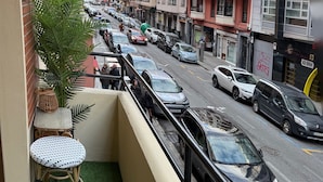 A balcony with a small table and chair, a potted plant, and a view of a busy street with parked cars and multi-story buildings. - APARTAMENTO  NUEVO, LUJOSO, CENTRO DE BILBAO. (Bilbo)