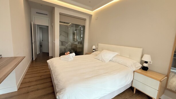 A hotel room with a bed, bedside tables, a nightstand, a wooden floor, a bathroom with a glass shower, and a hallway leading to another room. - APARTAMENTO  NUEVO, LUJOSO, CENTRO DE BILBAO. (Bilbo)