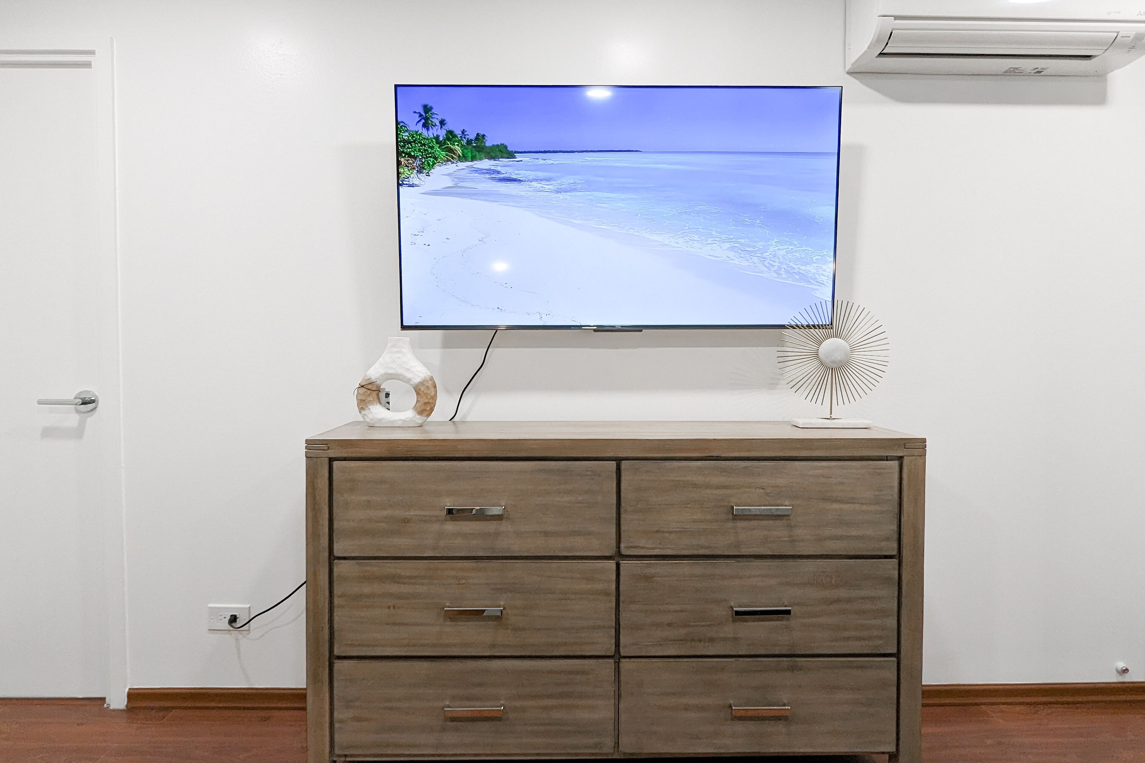 A room with a TV on a wooden cabinet, a wall-mounted air conditioner, and a power outlet.