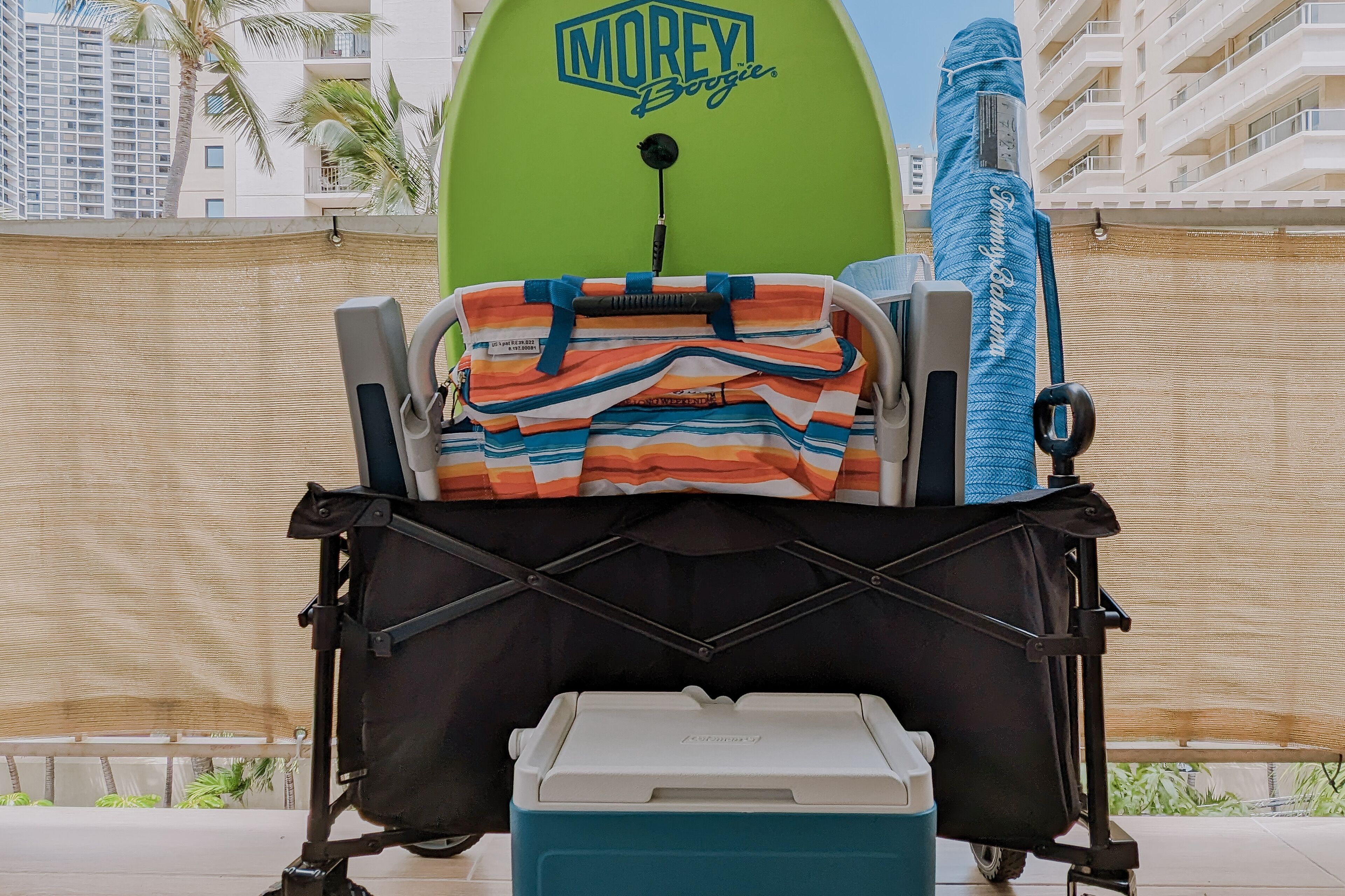 Una silla de playa con una toalla colorida y una toalla azul con el texto "Morey Boogie", una tabla de surf verde y un enfriador en un balcón.