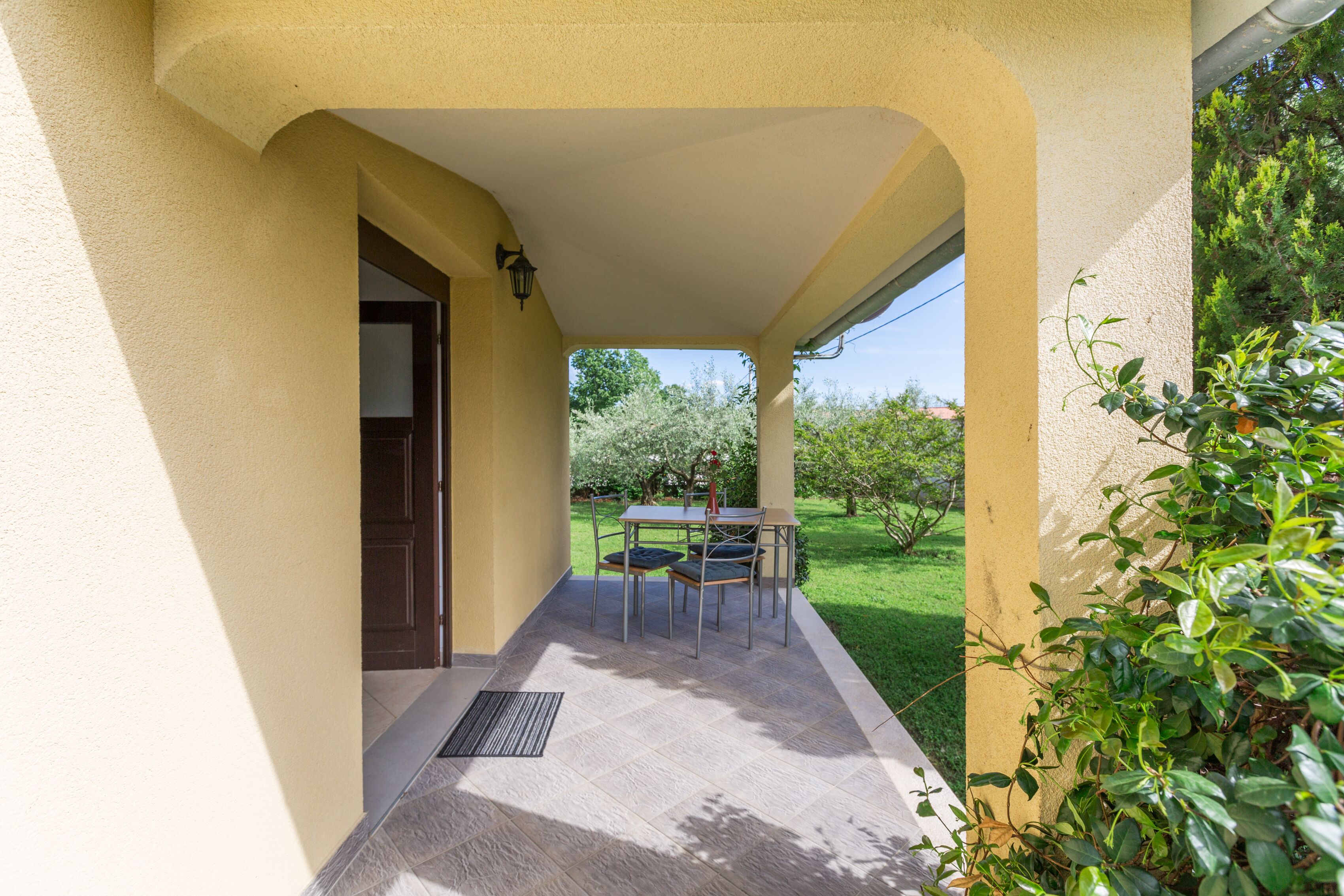 Un patio techado al aire libre con mesa y sillas, una puerta y vistas a un jardín.