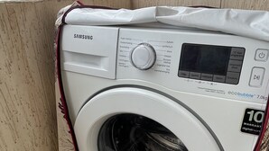 Une machine à laver Samsung dans une buanderie, avec un sol carrelé et une vue sur un bâtiment en arrière-plan.