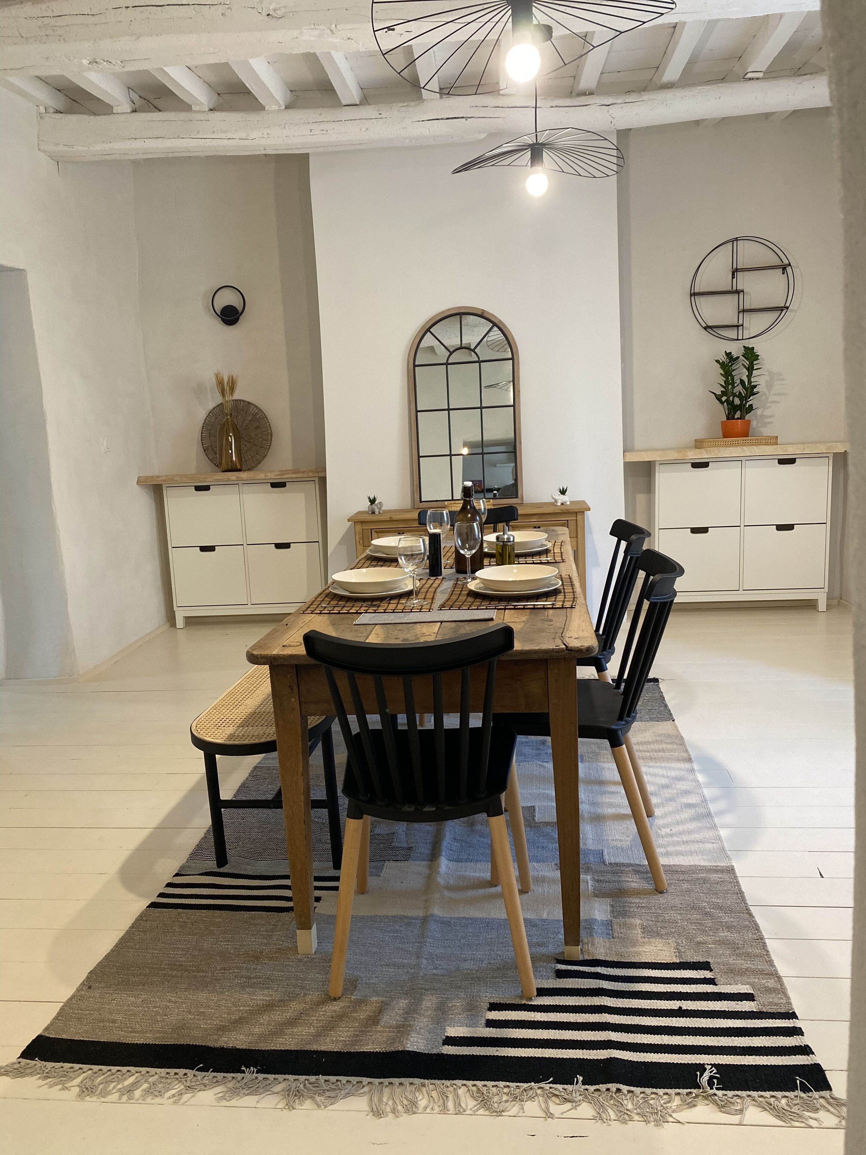 Un comedor con mesa de madera, sillas negras, alfombra estampada y un mueble blanco con espejo.