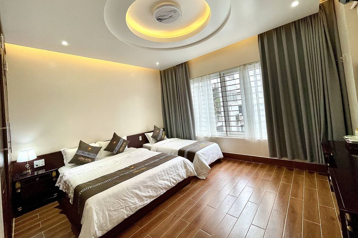 Habitación de hotel con dos camas, un ventanal grande con cortinas, suelo de madera y un diseño moderno en el techo.