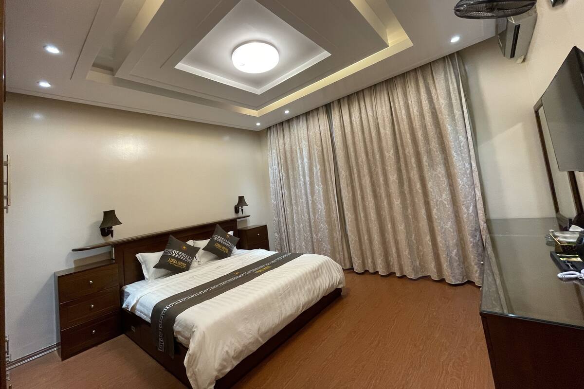 Una habitación de hotel con cama, mesita de noche, lámpara, luz de techo, espejo y ventana con cortinas.