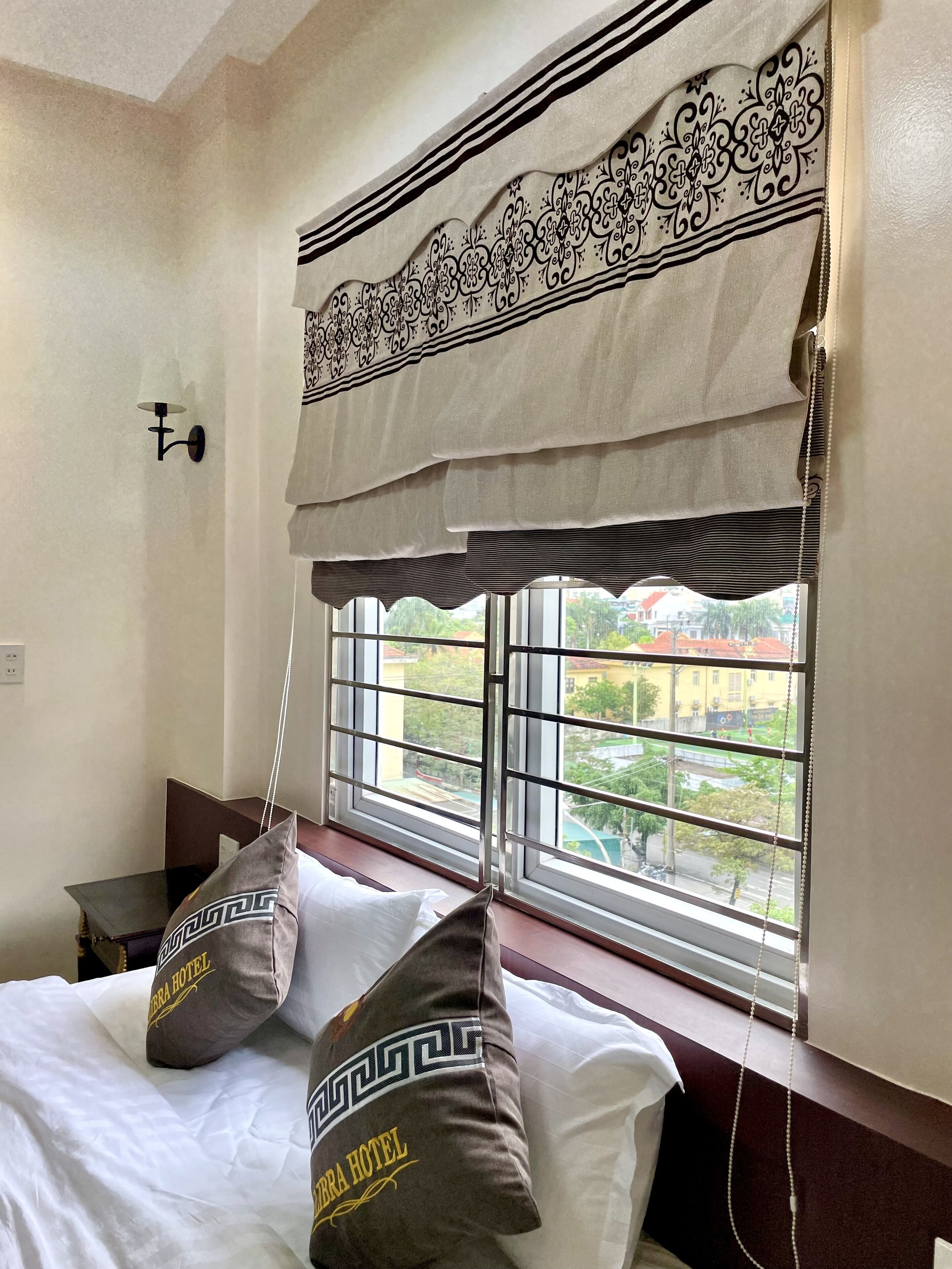 Ein Hotelzimmer mit einem Bett, dekorativen Kissen, einem Fenster mit Blick auf Gebäude und einer wandmontierten Lampe.