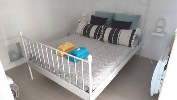 Una cama individual con estructura metálica blanca, ropa de cama gris estampada y dos almohadas a rayas negras y blancas.