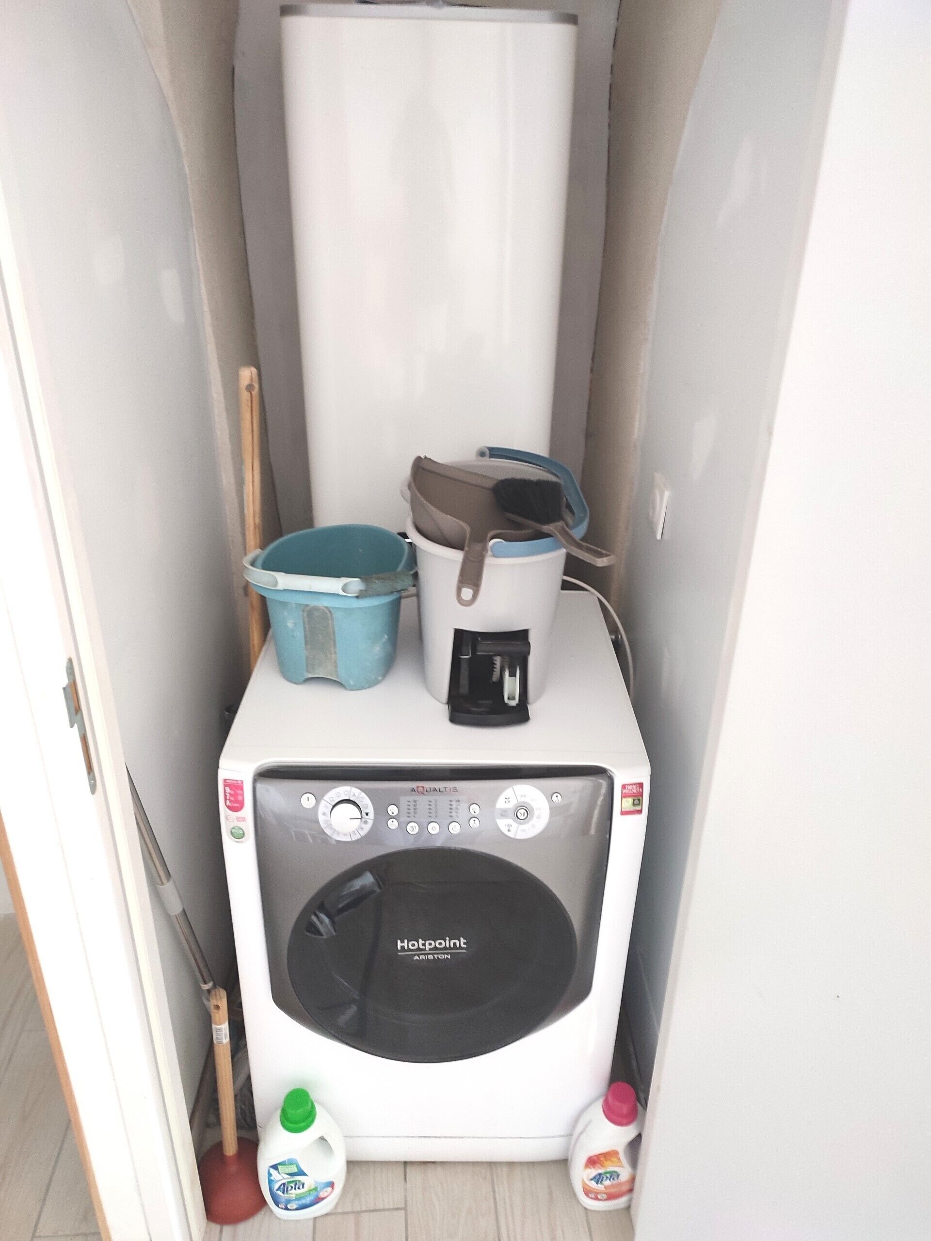 Een compacte, witte wasmachine met een klep aan de voorkant, een blauwe emmer, een borstel en schoonmaakmiddelen op een klein aanrecht.