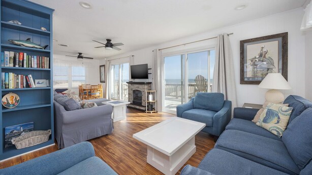 A living room with blue sofas, a white coffee table, a bookshelf with books and decorative items, a fireplace, and a large window with a view. - 5852 - Lega Sea - Oceanfront Cottage | Pool, Views & Hot Tub (Nags Head)