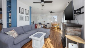 A living room with a grey sofa, white coffee table, and a fireplace. There are ceiling fans, a TV mounted on the wall, and a bookshelf. - 5852 - Lega Sea - Oceanfront Cottage | Pool, Views & Hot Tub (Nags Head)