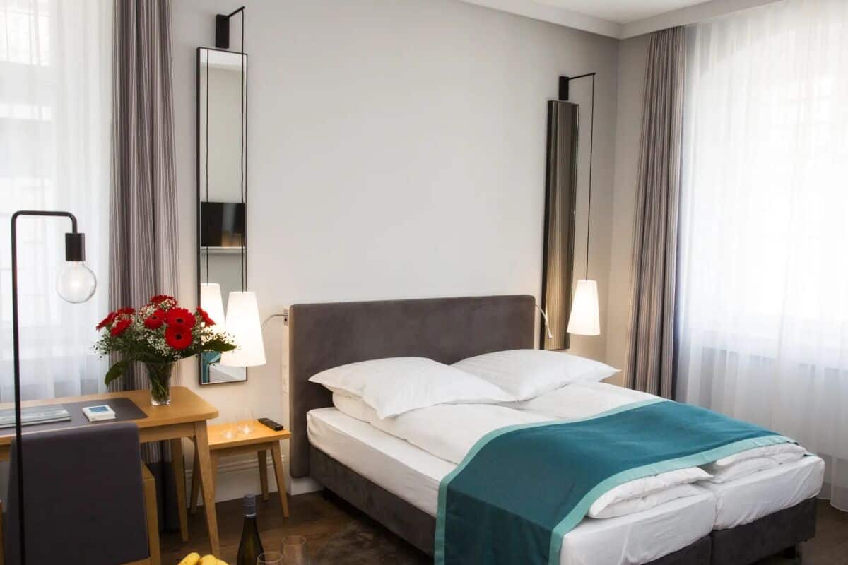 A hotel room with a bed, bedside table, lamp, and a small round table with fruit and a glass of wine.