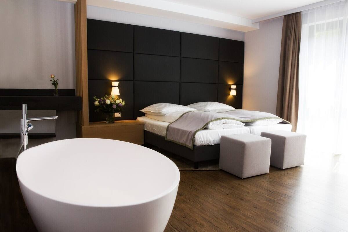 A modern hotel room with a large white bathtub, a dark headboard, and a wooden floor.