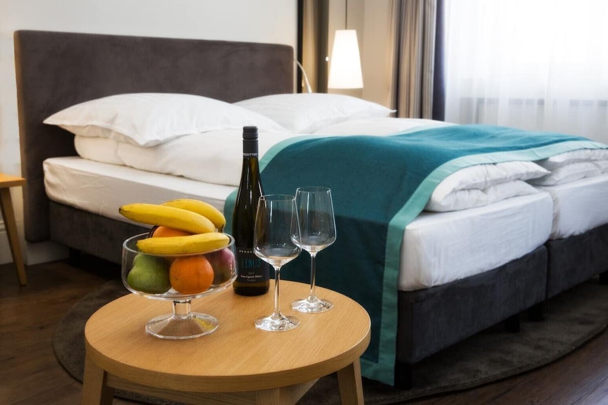 A hotel room with a bed, a round wooden table with a fruit bowl, a bottle of wine, and two wine glasses.