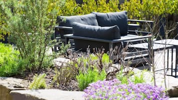 Ein Außenbereich mit Sitzgelegenheiten, darunter eine Couch und Stühle, umgeben von verschiedenen Pflanzen und Blumen.