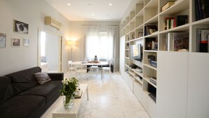 Ein modernes Wohnzimmer mit einem dunklen Sofa, einem weißen Couchtisch mit einer Vase, einem Essbereich mit Tisch und Stühlen sowie einem großen Bücherregal, gefüllt mit Büchern.
