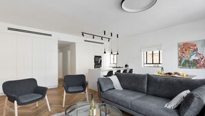 A modern living room with a grey sofa, two armchairs, a glass coffee table, and a kitchen in the background. - Spacious & Modern 3BD Apartment 2-min From Beach (Tel Aviv)