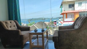A room with two brown armchairs and a wooden coffee table, a large window with a view of a body of water and buildings. - Hotel De Pebbles (Port Blair)