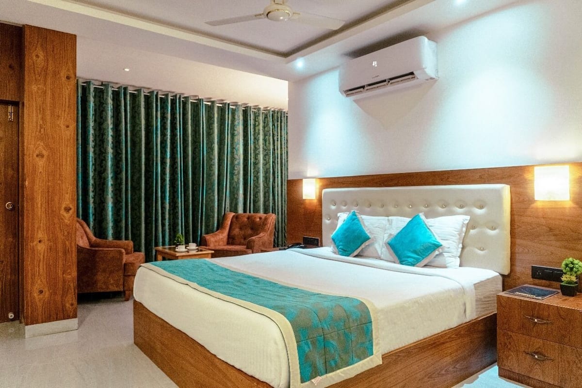 A hotel room with a large bed, a wooden wardrobe, a seating area with a chair, and an air conditioning unit on the wall.