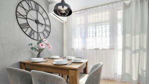 Un comedor con una mesa de madera dispuesta para seis personas, rodeada de sillas grises, un reloj de pared grande y un jarrón con flores.