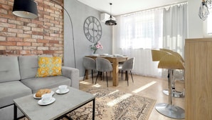 Una sala moderna con un sofá gris, una mesa de comedor con sillas, una mesa de centro con tazas y un pastel, una pared de ladrillo y un reloj grande.