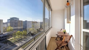Un balcón con mesa y sillas, ventanas amplias que ofrecen vista a la ciudad y una lámpara colgante.