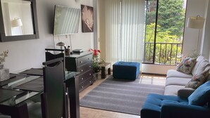 Un salon moderne avec une table à manger en verre, un canapé et un pouf bleu. Un téléviseur est fixé au mur, il y a une fenêtre donnant sur un espace verdoyant et une lampe sur une table d’appoint.