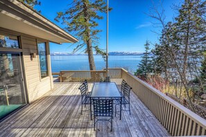 Terraza de madera con una mesa y sillas, con vista a un lago y montañas al fondo.
