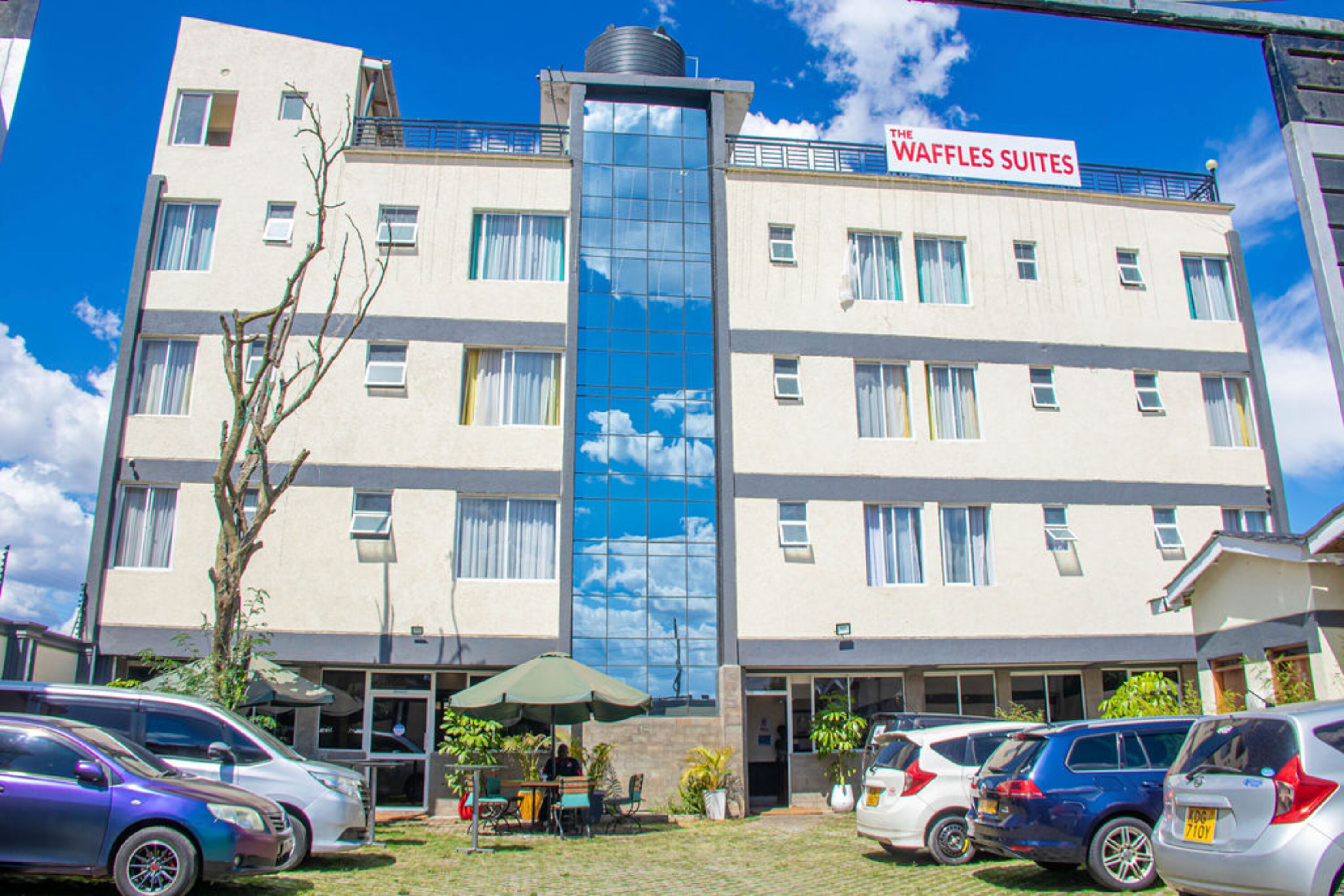 A multi-story hotel with a sign reading "The Waffles Suites." There are several cars parked in front, and a small outdoor seating area with umbrellas.