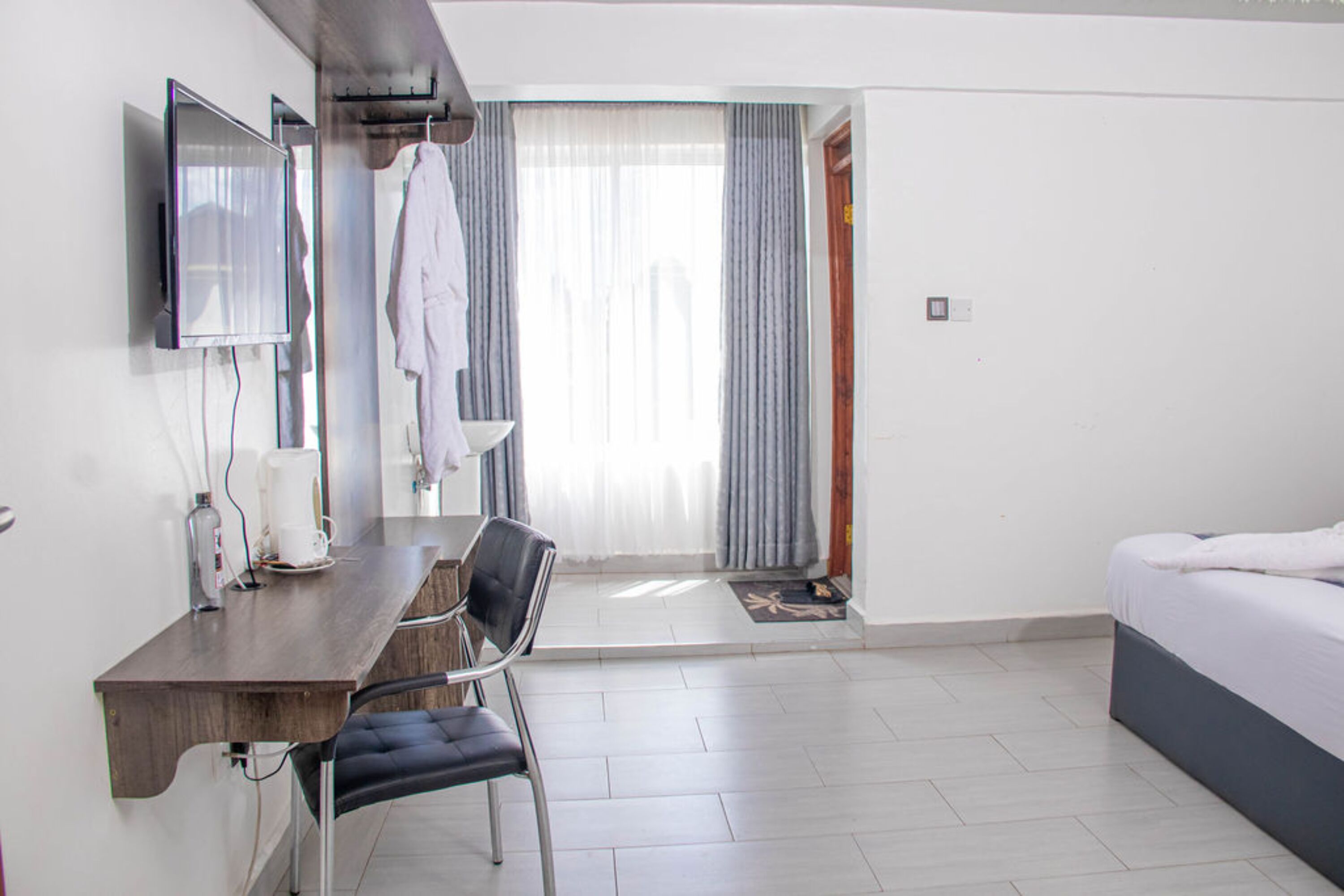 A hotel room with a bed, a desk with a chair, a TV mounted on the wall, and a window with curtains.
