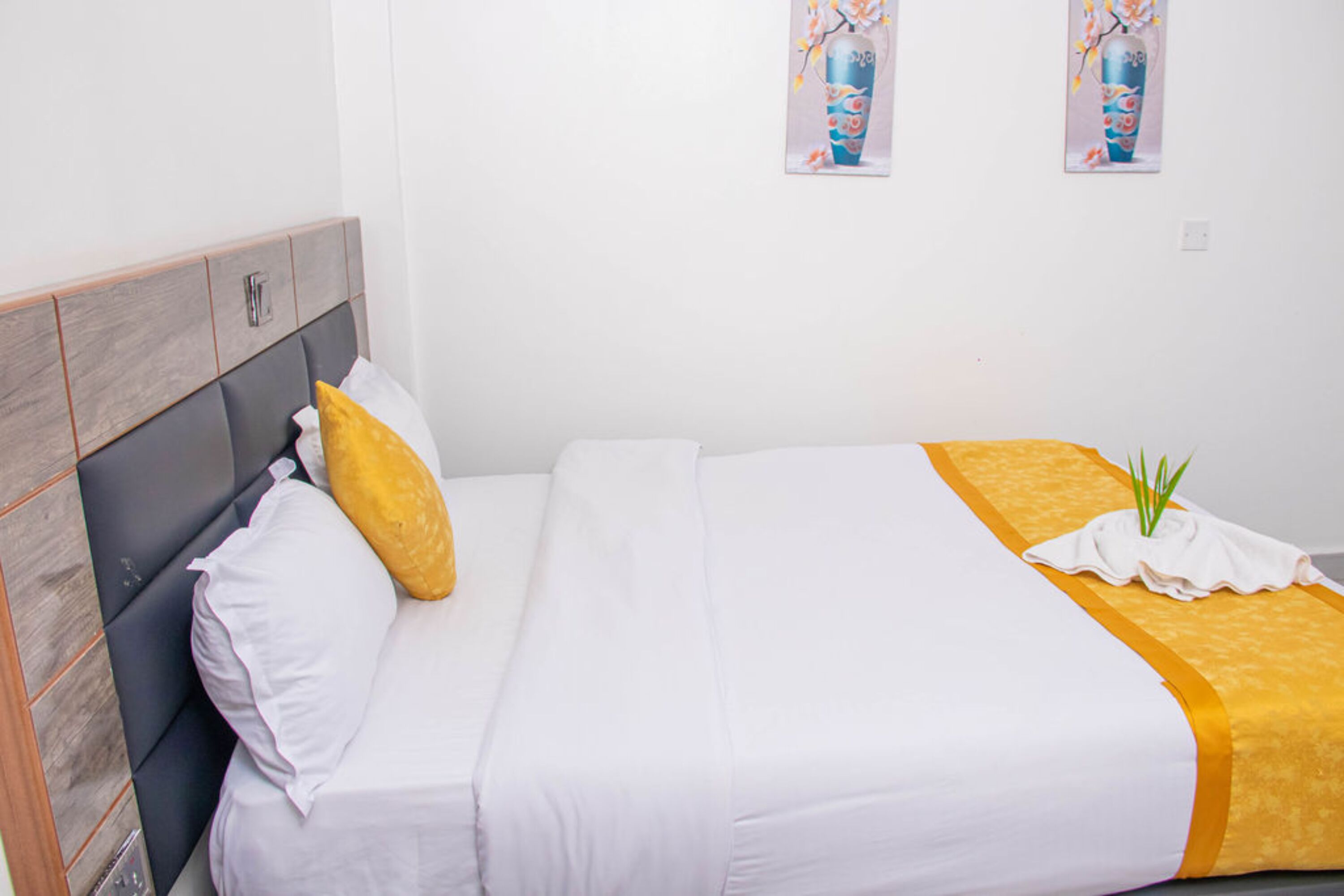 A neatly made bed with white and yellow bedding, a grey headboard, and two framed artworks on the wall.