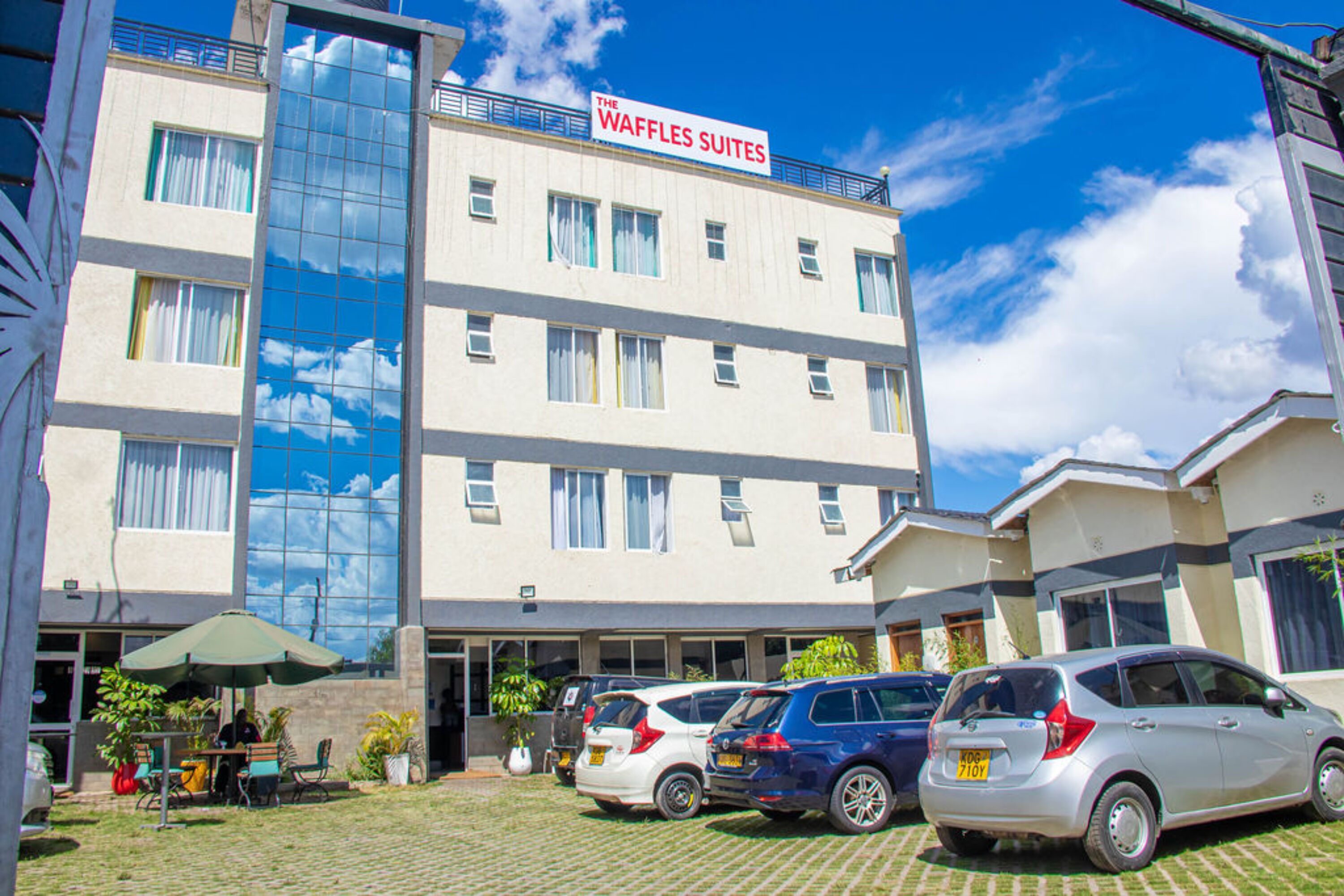 A hotel named "The Waffles Suites" with a modern design, featuring a large glass facade, a parking area with several cars, and outdoor seating with umbrellas.