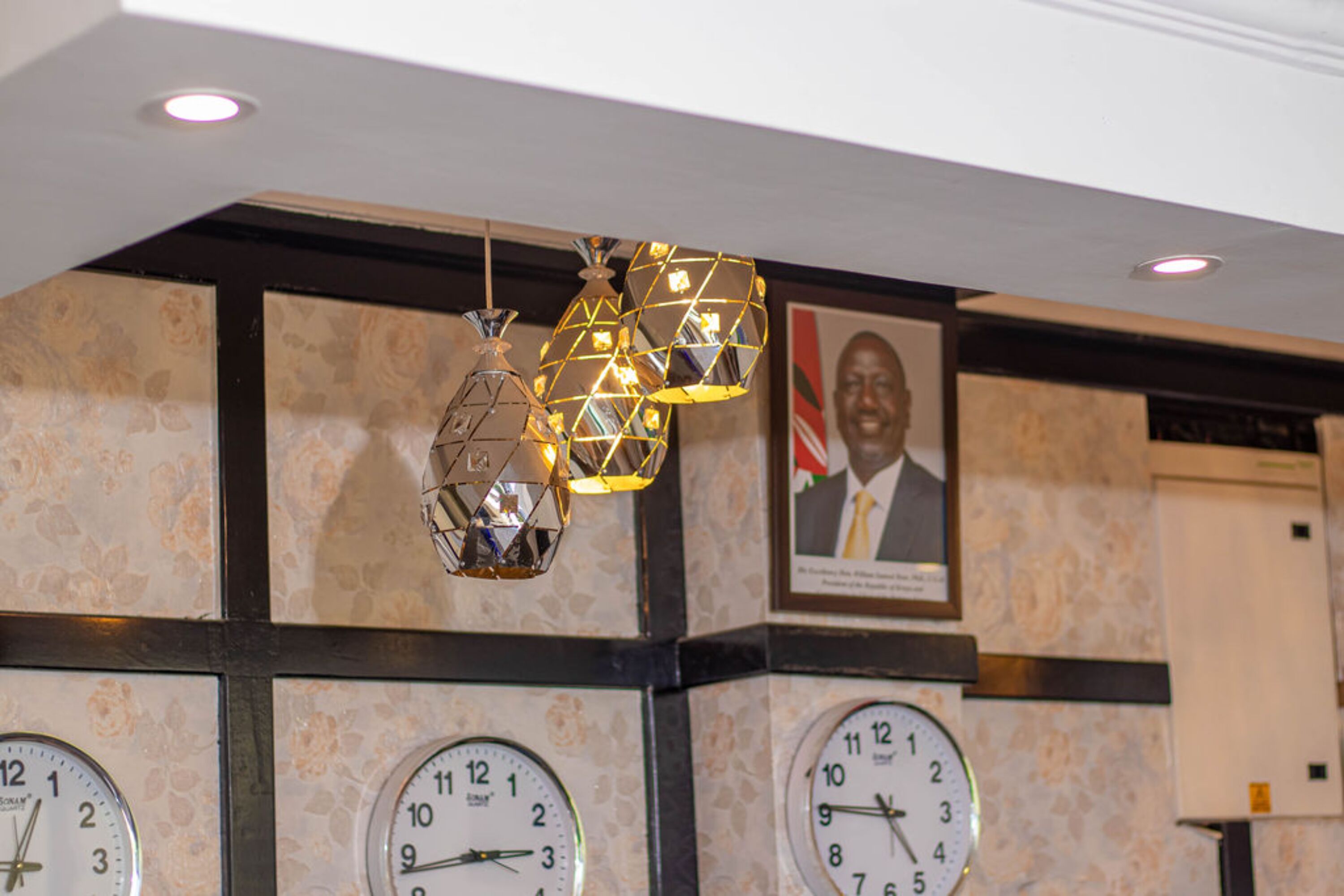 A wall with three clocks, a framed portrait, and hanging light fixtures.
