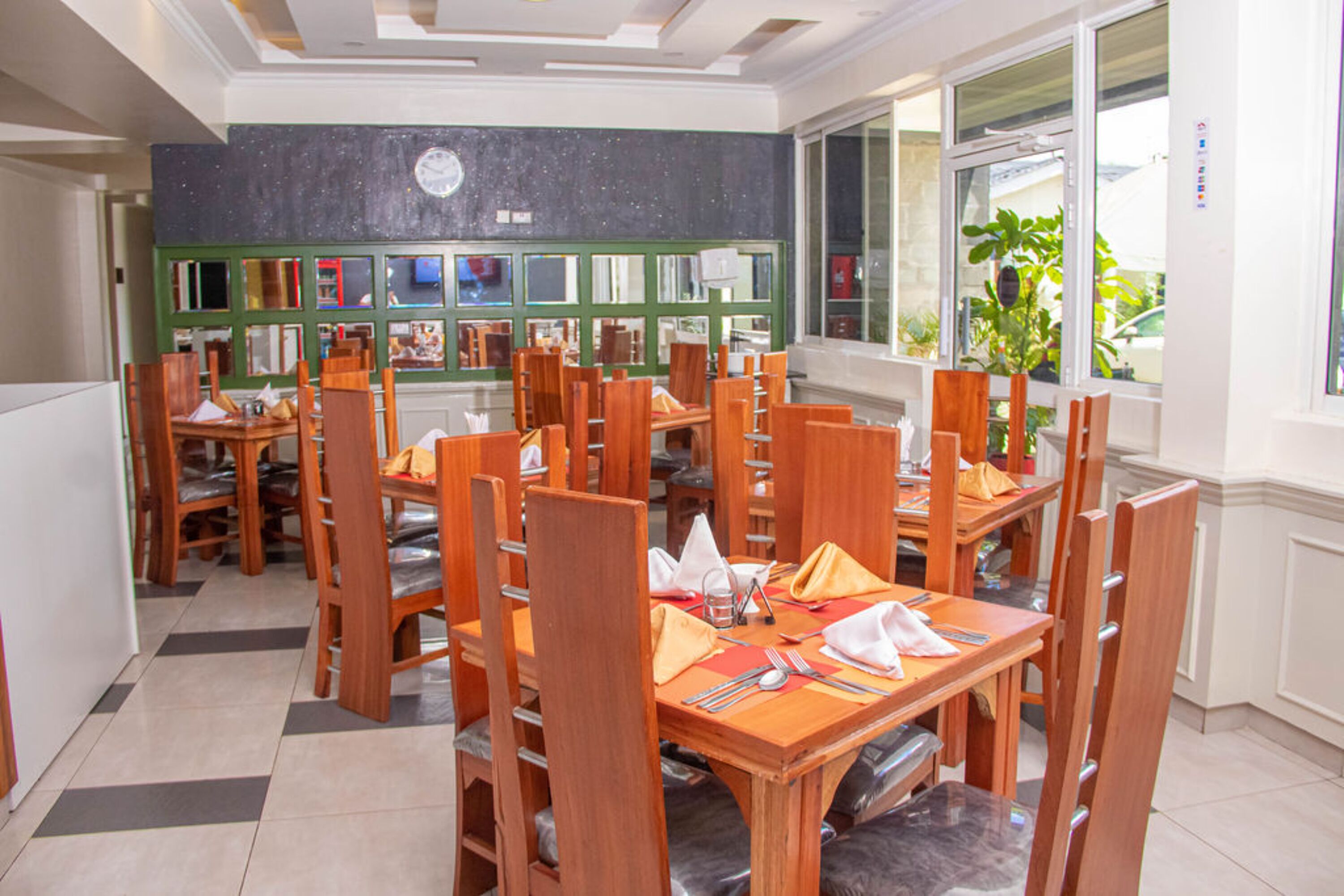 A dining area with multiple wooden tables and chairs, a green wall with framed pictures, and a window with a view of greenery outside.