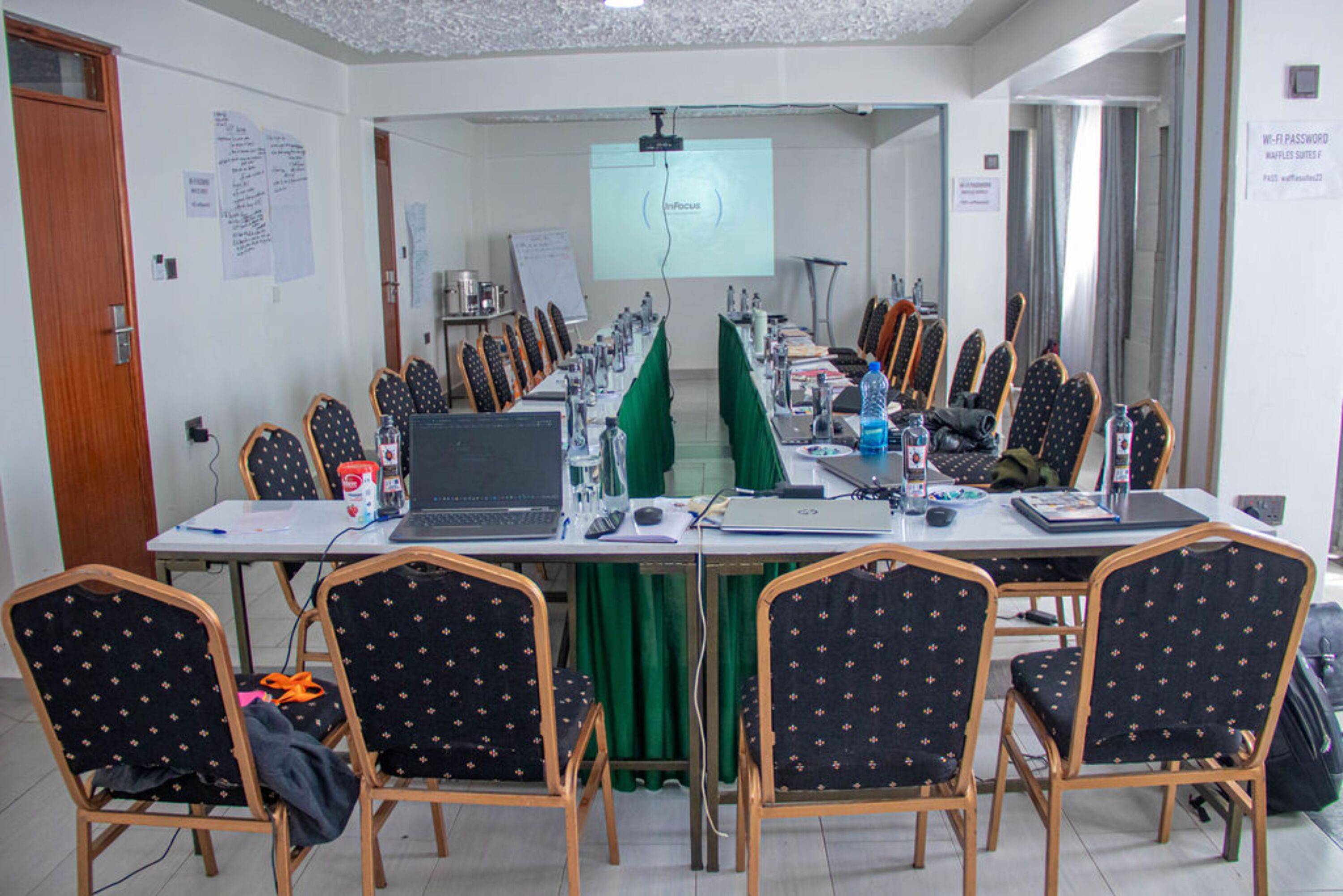Un salón de conferencias con una larga mesa, sillas, computadoras portátiles y una pantalla para proyector.