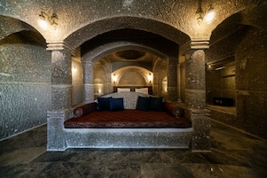 Habitación de paredes de piedra con una cama bajo un techo abovedado, columnas de piedra y luces fijadas a la pared.
