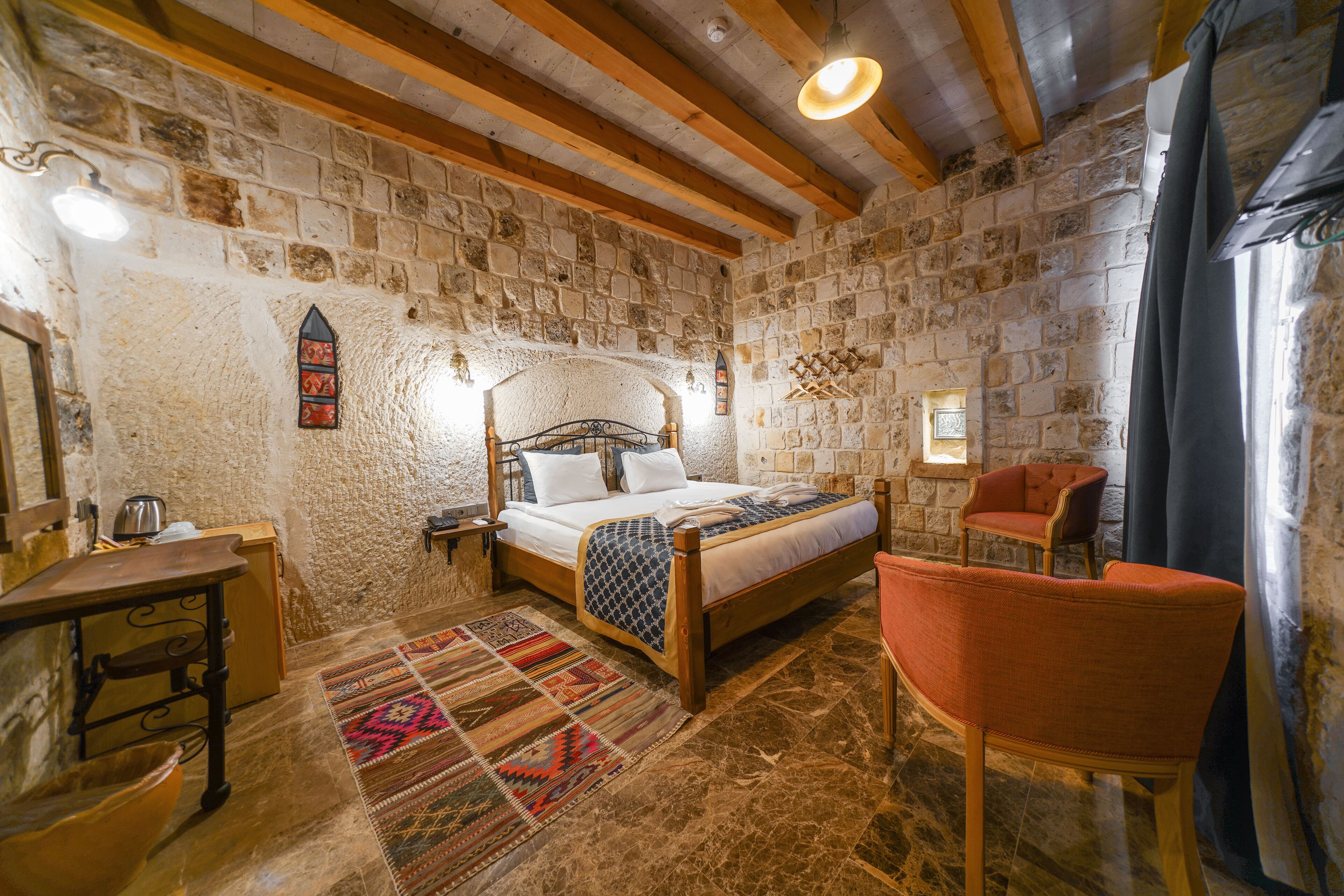 Una habitación rústica con pared de piedra, vigas de madera, una cama con colcha estampada, una silla y una mesita con hervidor.