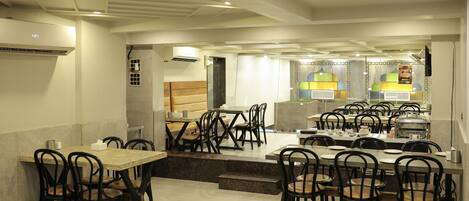 A restaurant with multiple tables and chairs, a kitchen area in the background, and an air conditioning unit on the wall.